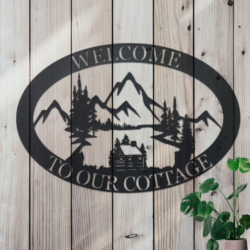 Metal oval sign of a cabin surrounded by mountains and trees. With Welcome to our Cottage on the border of the sign.