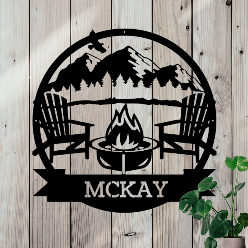 Round metal sign with 2 chairs around a campfire and mountains and trees in the the background. Custom name at the bottom. 