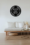 Black circular metal sign with the words bless our nest written in the middle hung above a white couch. 