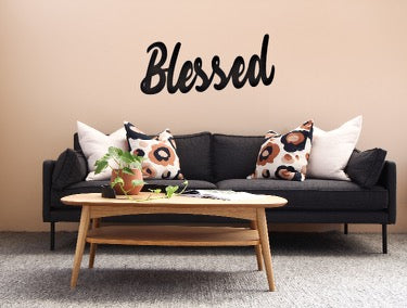 Metal sign with the word Blessed in cursive writing hung on a wall above a couch with a coffee table in front of couch.