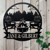 Round black metal sign with 2 muskoka chairs around a bonfire and a row of pine trees behind them. Banner at the bottom of the sign for a custom name.