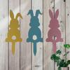 Three bunny garden stakes. One painted yellow with one ear up and one ear down. One painted blue with both ears down. One painted pink with both ears up.